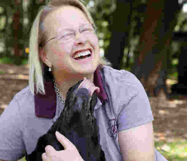 A person laughs as their black Labrador Guide Dog licks their face.