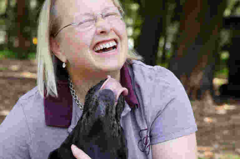 A person laughs as their black Labrador Guide Dog licks their face.