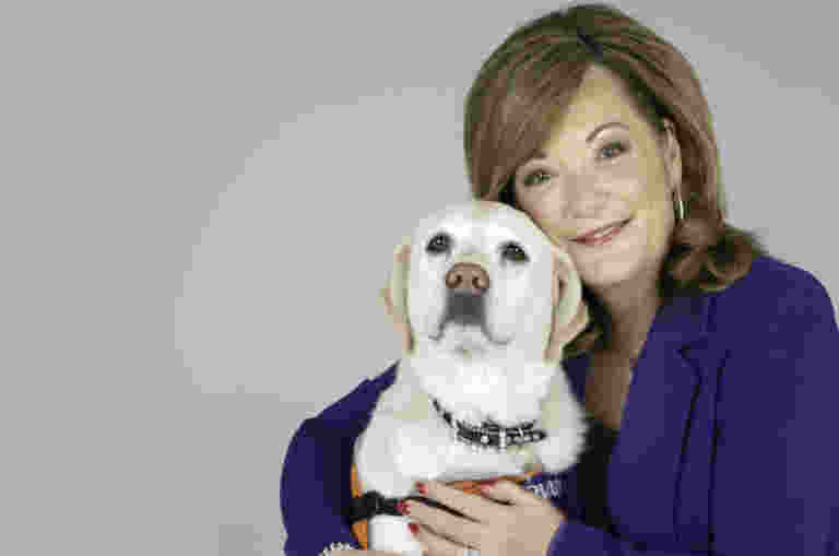 Karen Hayes is standing next to her yellow Ambassadog Willow. She has her arms around her and is smiling at the camera.