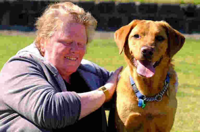 Marg is sitting in a park on grass next to her Guide Dog Scottie. She is leaning towards him with her arm on his back. Scottie is looking directly at the camera and smiling