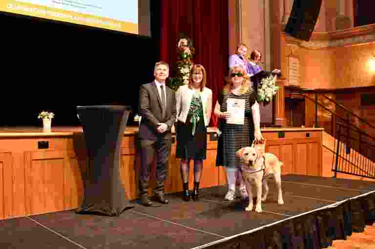 On stage at the Hawthorn Town Hall on Graduation Day. Pictured is a Guide Dogs client with her yellow Guide Dog standing beside GM of Vision Services Leah McFadzean and Chairperson Iain Edwards