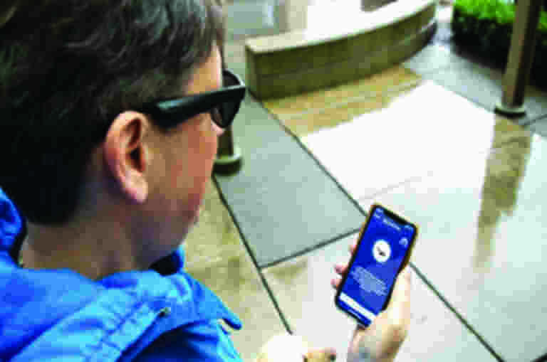 A person is standing on the sidewalk with a yellow labrador next to them. They are looking at their smartphone