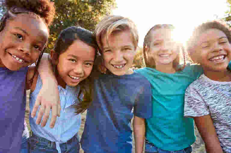 A group of 5 smiling kids are standing close together with their arms around each other smiling. They are outdoors and the sun is shining.