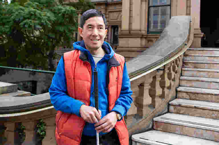 A Guide Dogs client is standing in front of a large stone staircase holding the handle of their white cane