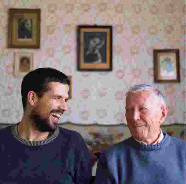 Two people, one adult & one older person, sitting on a couch looking at each other laughing.
