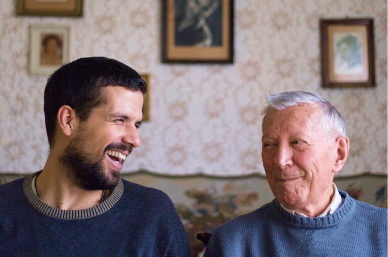 Two people, one adult & one older person, sitting on a couch looking at each other laughing.