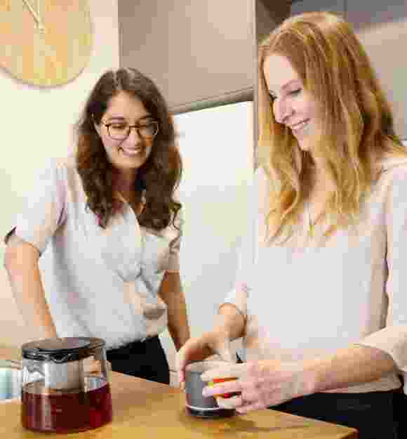Two people inside in the kitchen. One is making tea while the other is watching on.