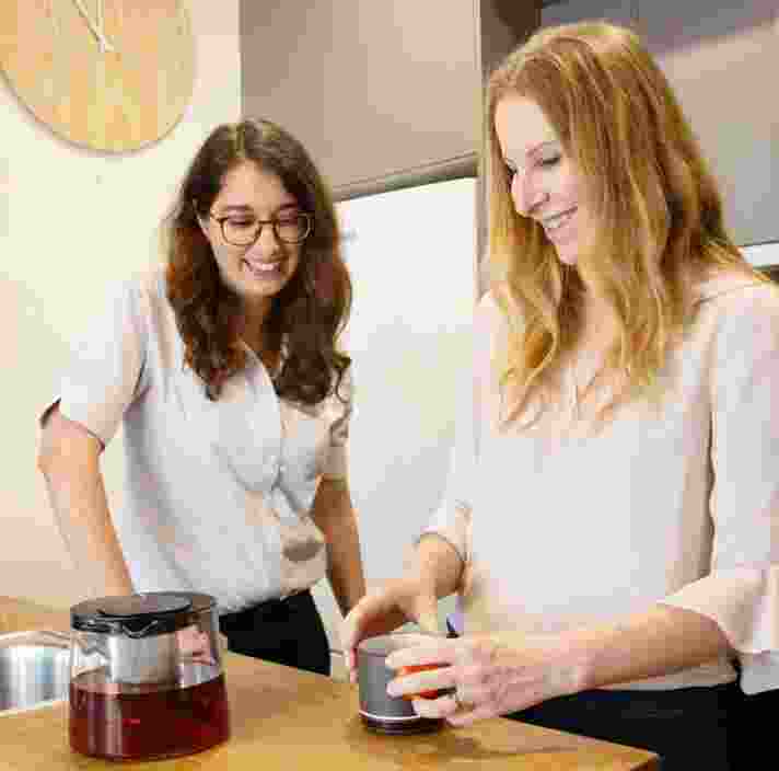 Two people inside in the kitchen. One is making tea while the other is watching on.