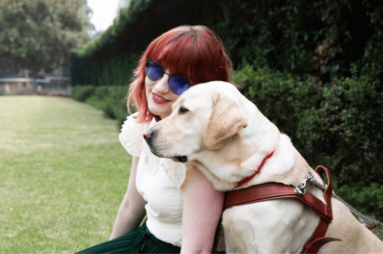 A person and a yellow Guide Dog, in harness, sitting on grass outside. The person is smiling at the camera and the Guide Dog is a profile view.