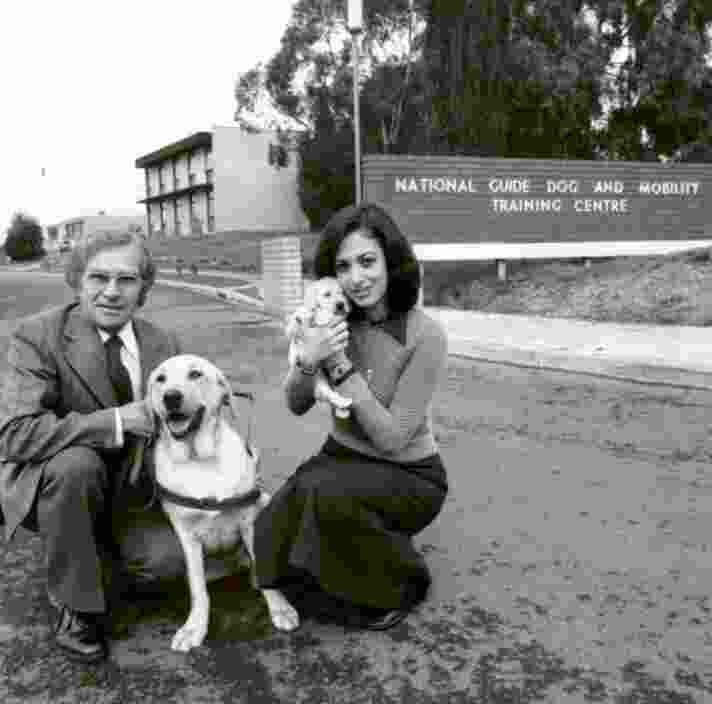 A 1940's black and white image of two people outside a Guide Dog Training Centre. One of the people in kneeling down next to a yellow labrador Guide Dog in harness and the other is holding a yellow ten week old labrador puppy. Both dogs and people are looking at the camera.