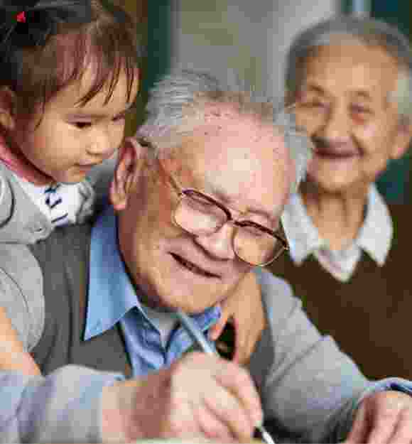 An older man writing while a young girl puts her arms around his shoulders, hugging him. The man is smiling and there is an older woman in the background who is not in focus but is also smiling.