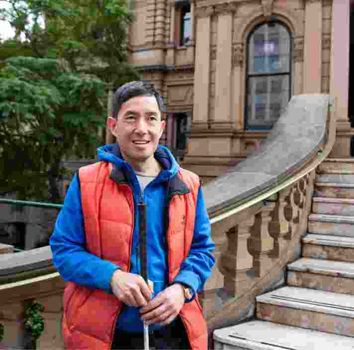 A person standing in front of a building holding their white cane. The person is smiling at the camera.