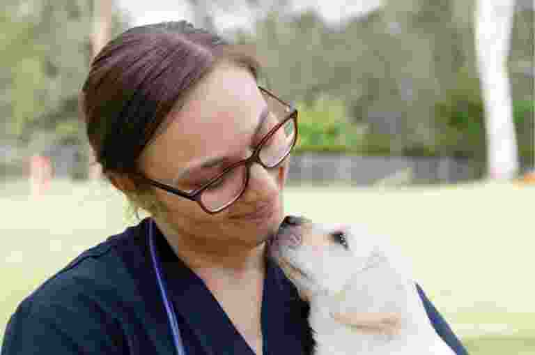 A veterinarian holding an eight week old yellow labrador puppy. The photo is taken outside and the puppy and vet are looking at each other, the veterinarian is smiling at the puppy.