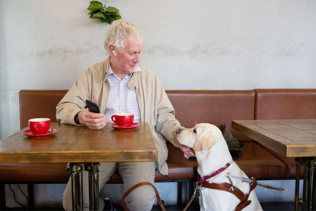 A person seated at a cafe table with a yellow labrador Guide Dog in harness seated next to their table. The person is reaching down touching the dogs face and the dog is looking up to the person.