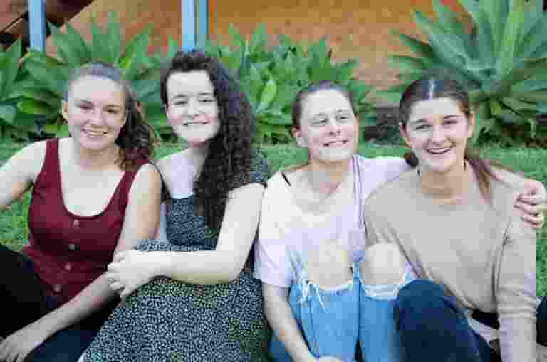 A group of four young adults sitting outside on the grass all in a row. Everyone is looking at the camera smiling.