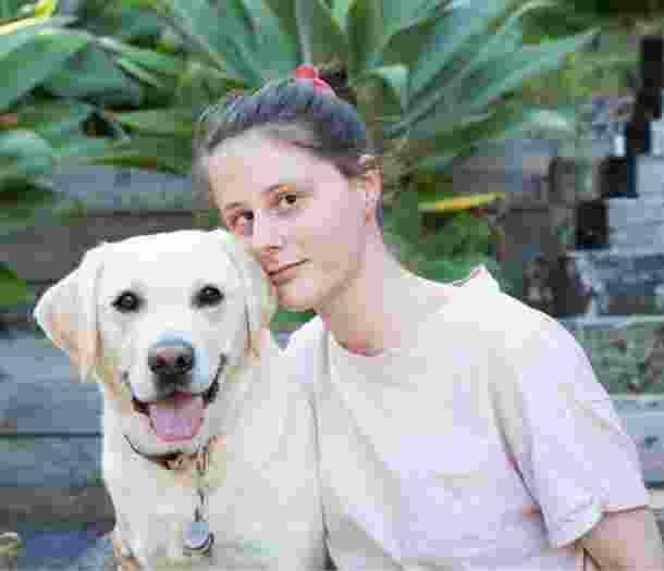 A person looking straight at the camera with their arm around a yellow labrador dog. They are seated at the bottom of a set of stairs.