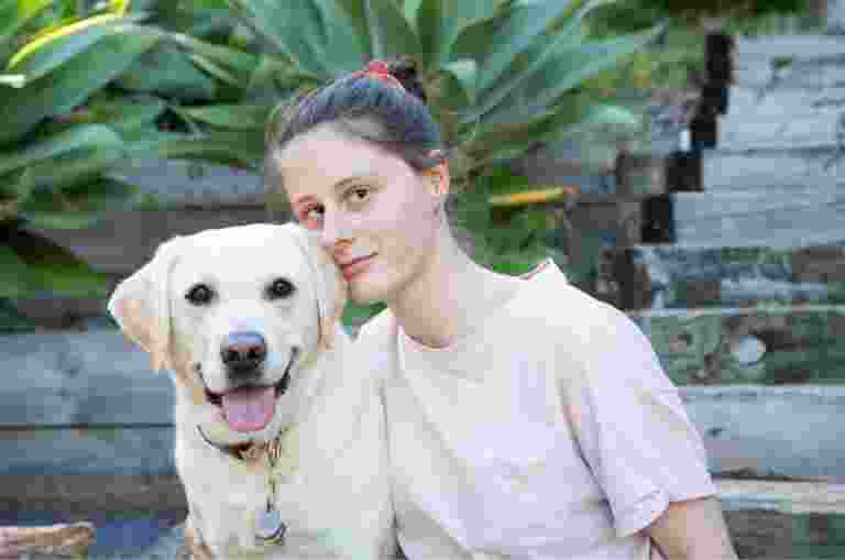 A person looking straight at the camera with their arm around a yellow labrador dog. They are seated at the bottom of a set of stairs.