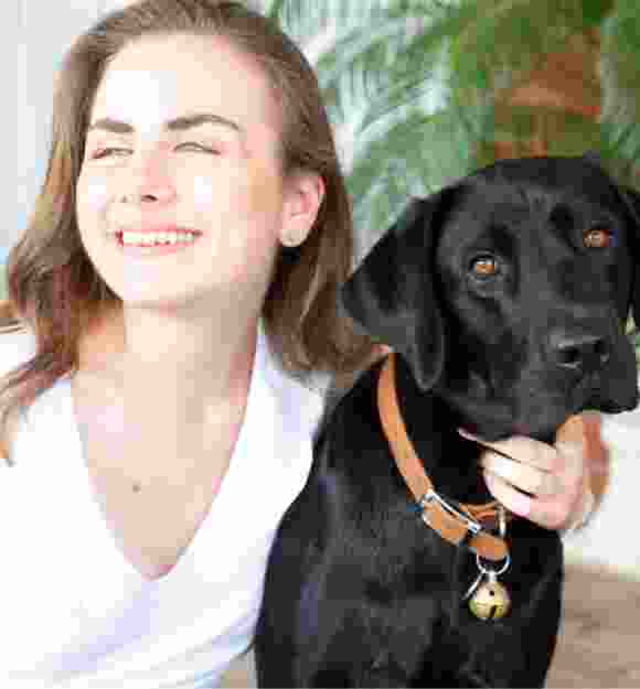 A person looking to the left of the camera smiling with their arm around the neck of a black labrador dog.