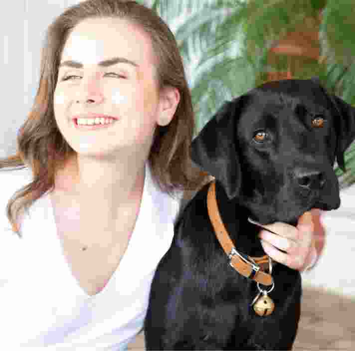 A person looking to the left of the camera smiling with their arm around the neck of a black labrador dog.