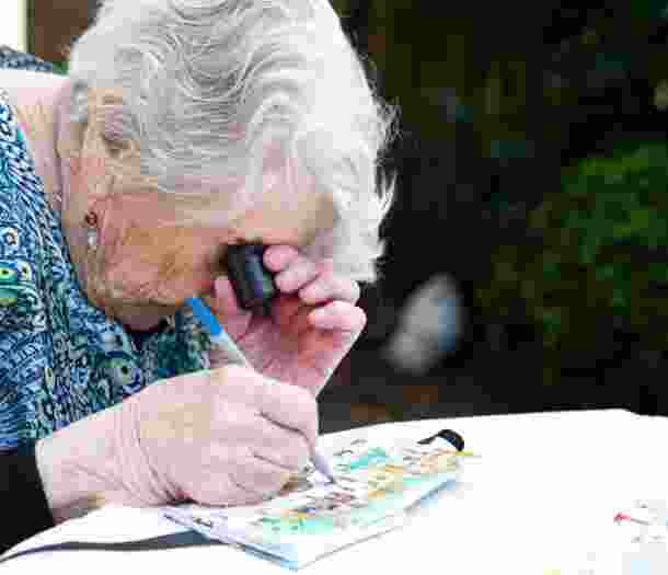 An older adult using a magnifier whilst painting, The person is painting onto a canvas.