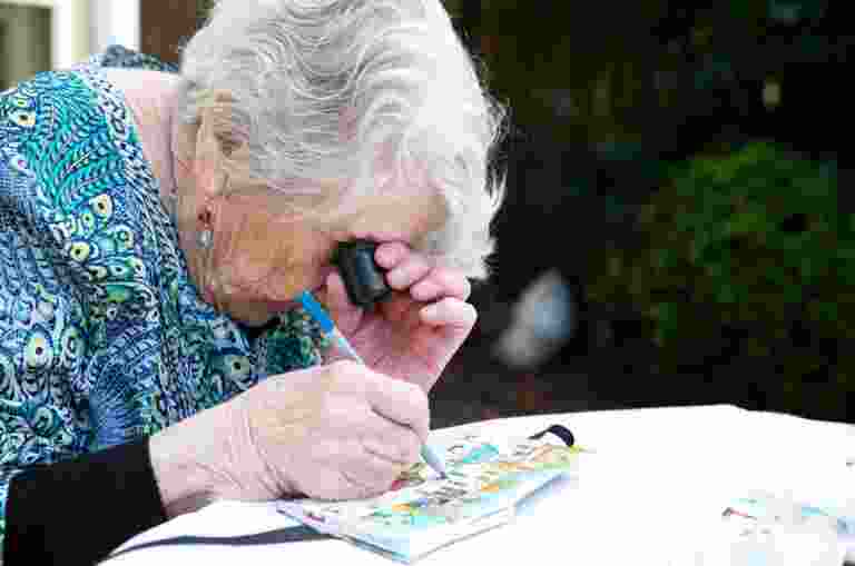 An older adult using a magnifier whilst painting, The person is painting onto a canvas.