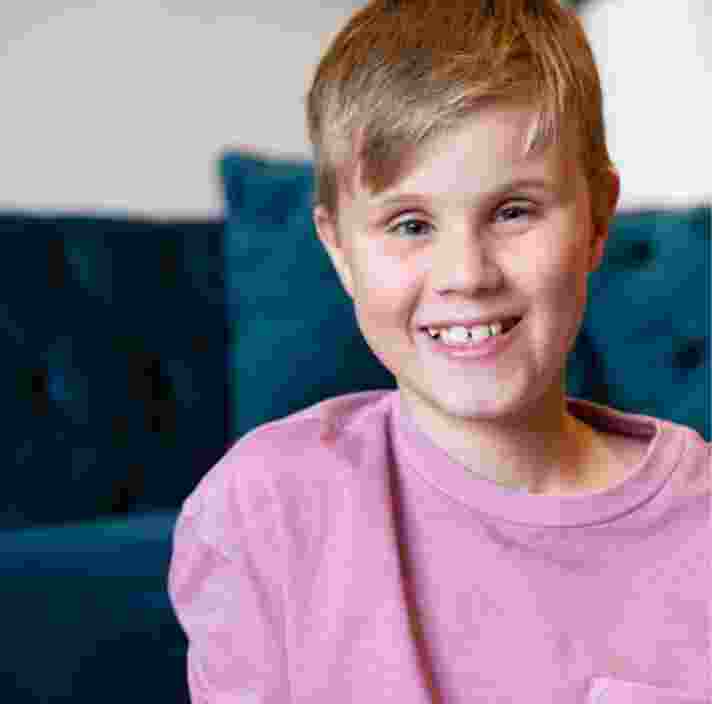 Close up of a young child with a big smile on their face. They are seated on the ground with a couch in the background.