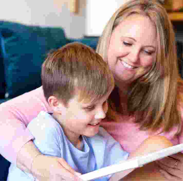 Young child sitting on the ground with their mother next to them. They are both smiling and looking at an Ipad.