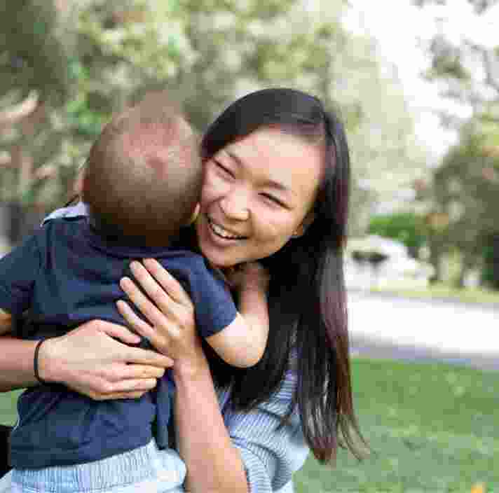 A mother and baby outside on the grass hugging each other. The mother is smiling and the baby has its back to the camera.