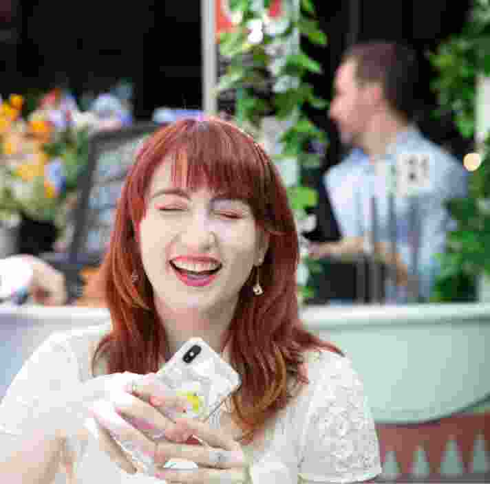 A person sitting at a cafe smiles and holds up their phone