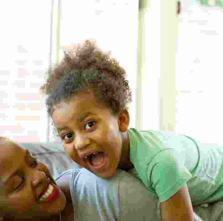 A mother and young child playing on the couch. The mother the child on top of her and the child has a big smile on their face.