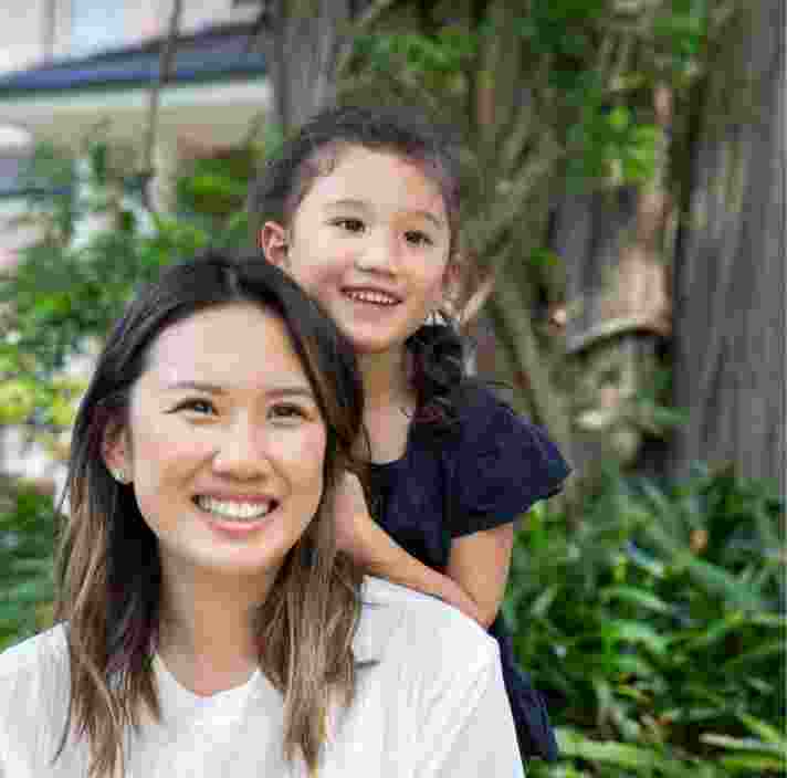 A mother and young child outside smiling at the camera. The young child is standing behind the mother with their hands on her back.