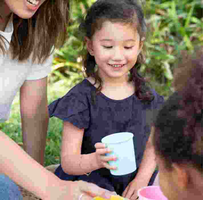 Two young children and a mother are playing with a tea set outside. The children and mother are smiling.