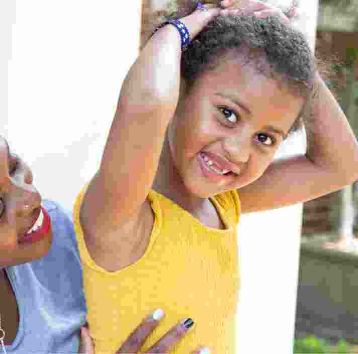 A young child smiling at the camera while their mother looks at them from behind. The mother is smiling at their child.
