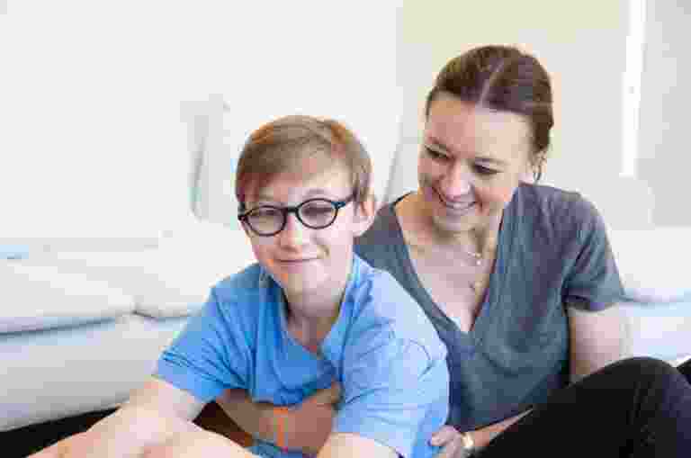 A young child with their mother seated on the ground. The mother is holding the child from behind and looking at her child. The child is looking at the camera smiling.