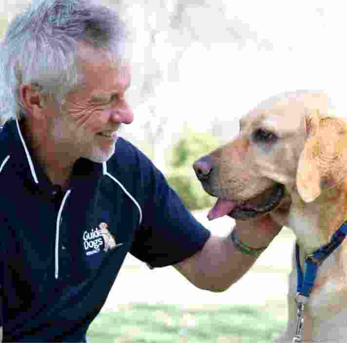 A Guide Dogs staff member and a yellow labrador dog sitting outside. The staff member is patting the dog whilst smiling and they are both looking at each other.