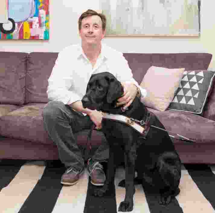 A person seated on a couch inside with a black labrador Guide Dog in harness next to them.
