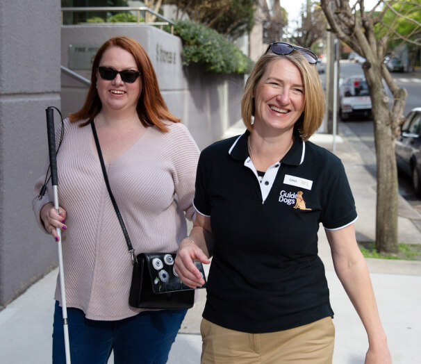 Two people walk together on the street, one holding a cane and one wearing a Guide Dogs polo shirt