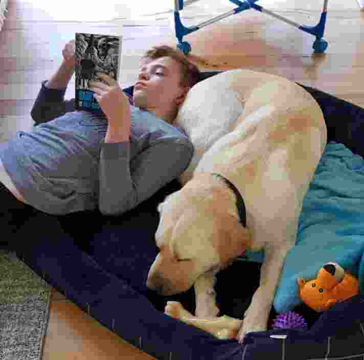 Max lies on the floor with his head resting on Bruno while he reads a book