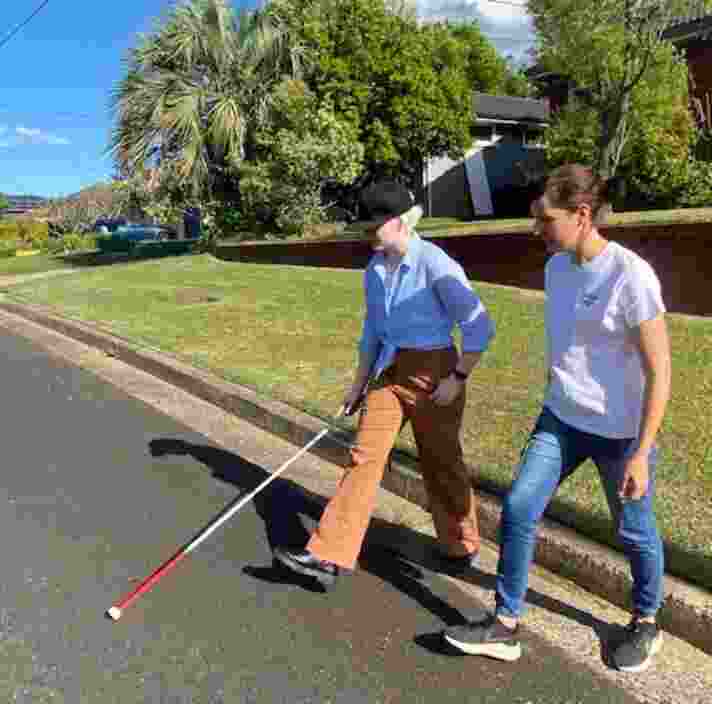 Zara crosses the road using her white cane next to her support worker Jen