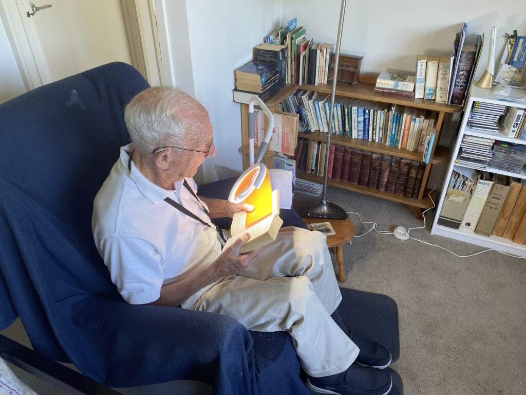 Dennis sitting in an armchair in his living room. He is reading a book using the magnifying lamp and a high contrast guide.