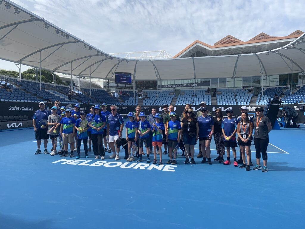 a group of children and adults all wearing blue and green shirts standing in the centre of a tennis court. 