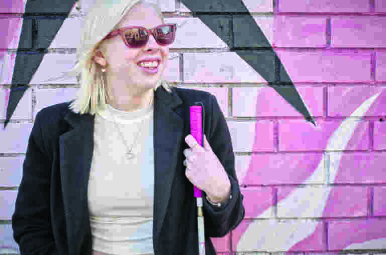 A young person with a pink cane standing in front of a pink wall.