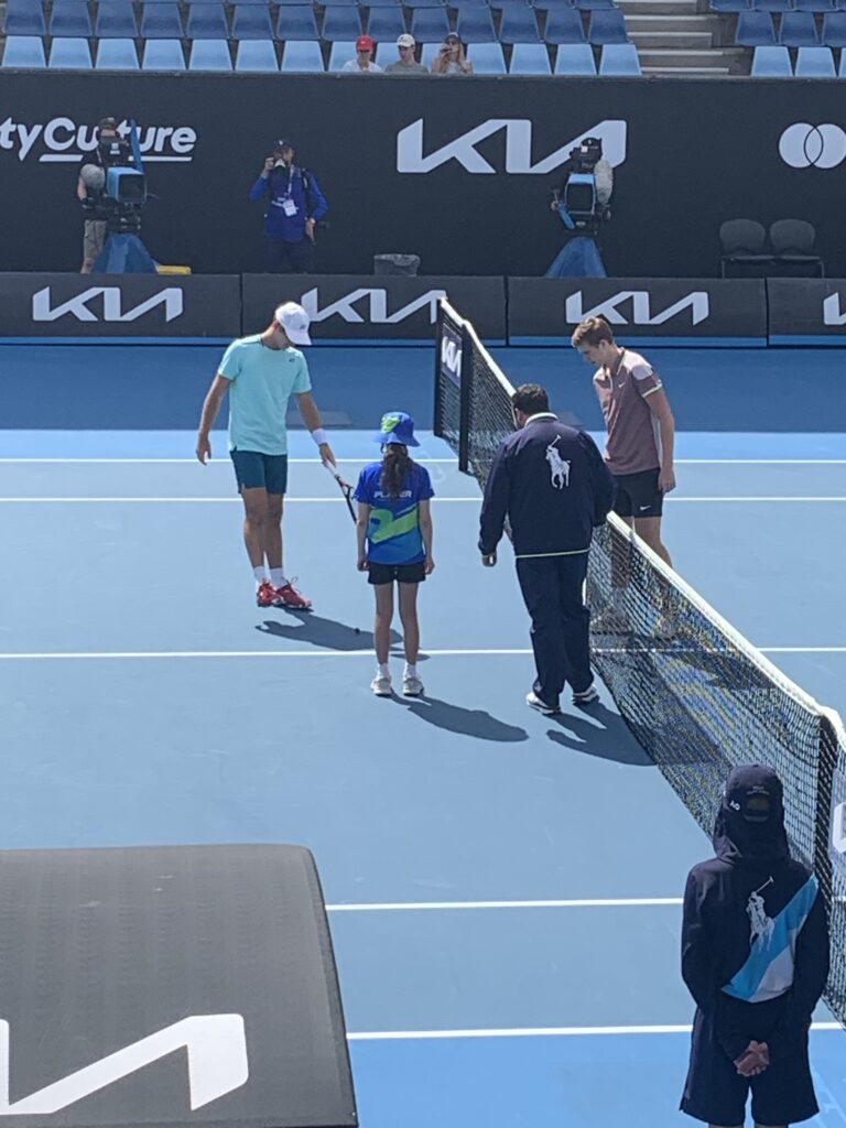 a young girl with a long brown ponytail tosses the coin on the blue tennis court with three male adults looking to see the result of the coin toss. 