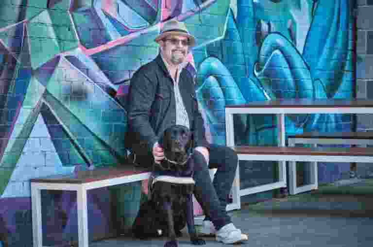 A person sitting in front of a blue graffiti wall, smiling, with their black Labrador Guide Dog sitting at their feet.