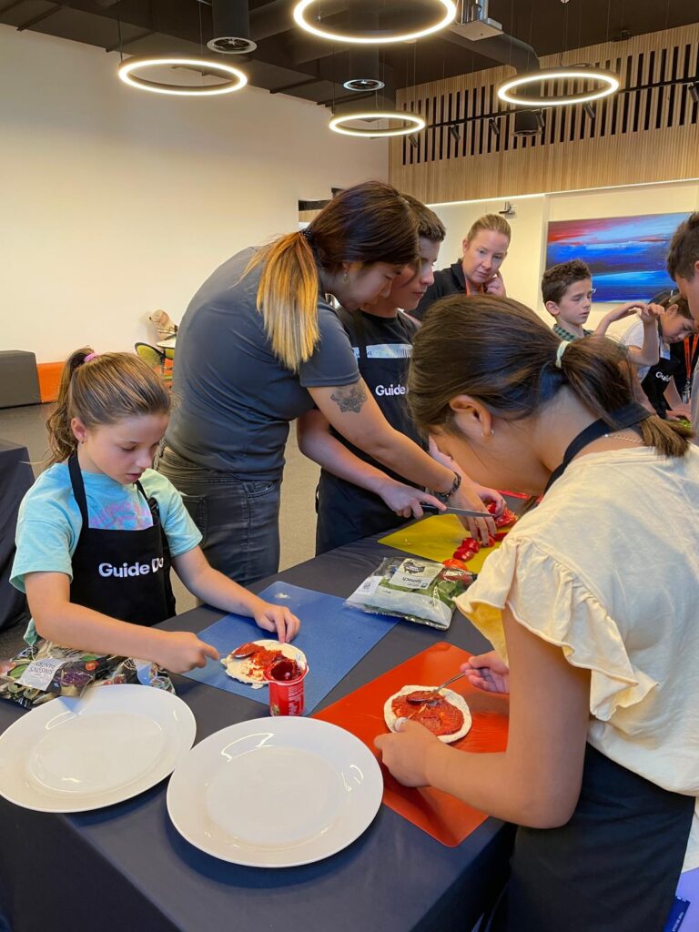A group of young people wearing black Guide Dogs branded aprons preparing food at a table, slicing tomatoes and spreading pizza sauce on small pizza bases. 