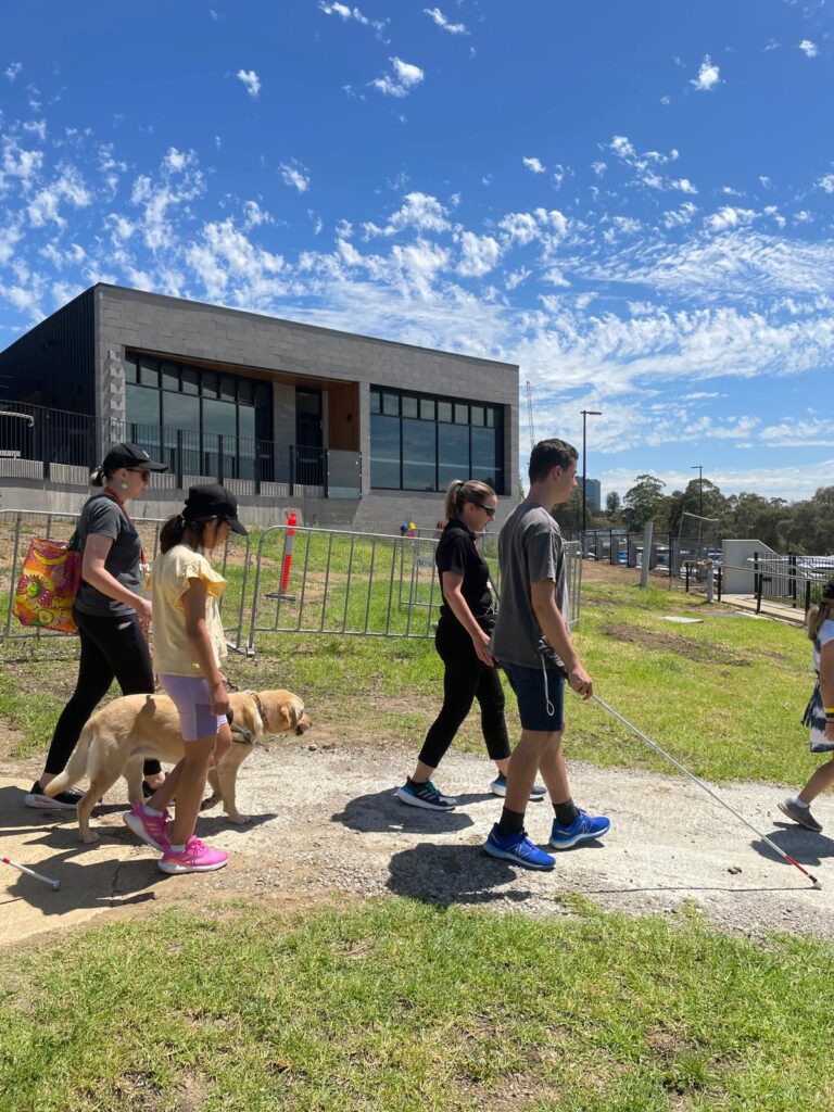A group of young people walk along a dirt track at Kew Campus. A young female walks with a golden Labrador Guide Dog with an instructor on the left-hand side of the dog, and a young man walks with a white cane with an instructor to his left. Grass, buildings and blue sky fill the background. 