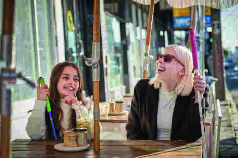 Two girls sit together at a table with their white canes, smiling and laughing.