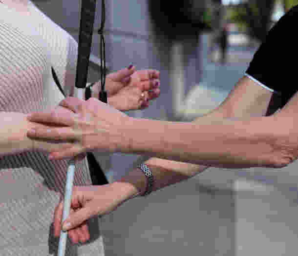 A close up image of a person holding a white cane. A Guide Dogs staff member goes to guide the Client.