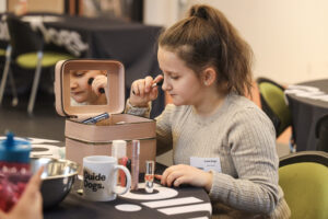 A young GDV Client looks into a pink makeup bag filled with products while holding a makeup brush to her face.