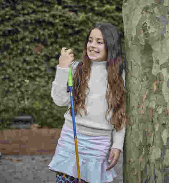 A young Client leaning against a tree, looking away smiling and holding onto her white cane.